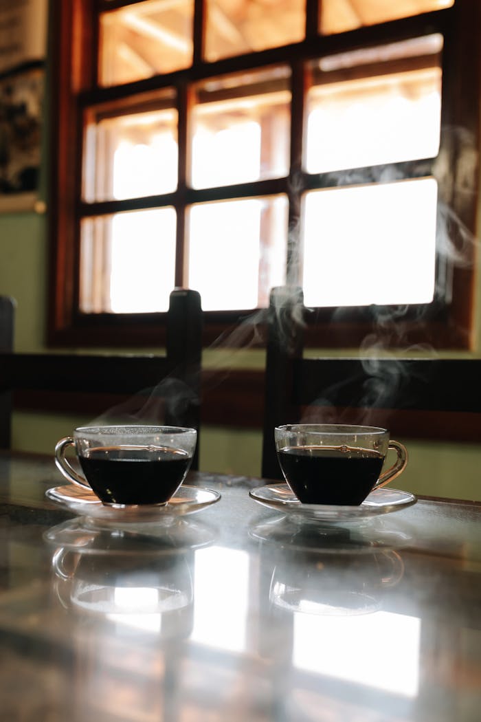 gallery-2 Warm, inviting steam rises from two cups of coffee in a rustic indoor café setting.